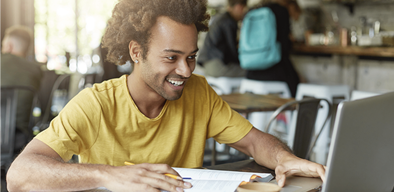 Rapaz jovem sorrindo e trabalhando em frente a um computador, com caneta na mão fazendo anotações